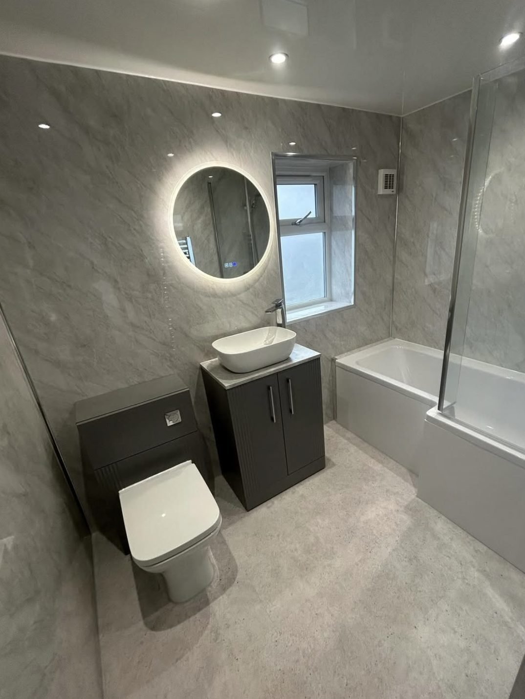 Grey marble effect panelled bathroom with backlit round mirror and bath screen